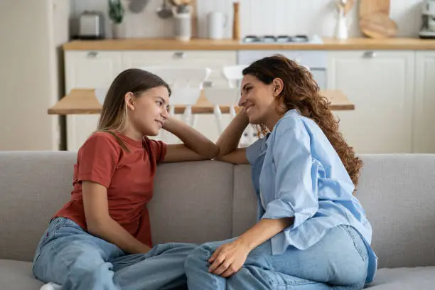 Loving mother listening to daughter with empathy and understanding while sitting together on sofa bonding at home, pre-teen girl child sharing secrets with mom, parent communicating with teenager