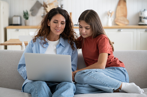 Young mother and teen girl daughter using laptop planning vacation, booking hotel or buying tickets while spending time together at home. Modern family mom and kid talk on video call with relatives