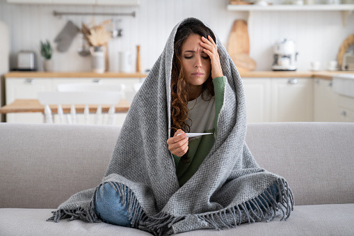 Sick exhausted woman with thermometer to measure body temperature sit on sofa at home wrapped in blanket. Suffering depressed girl experiences health problems due to abnormally cold winter weather