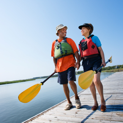 African American middle-aged couple walking on boat dock holding hands and carrying boat paddles.