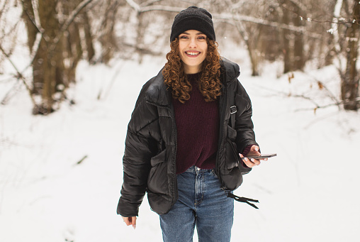 Redhead curly woman in black jacket and hat holding mobile phone in hand in winter forest.