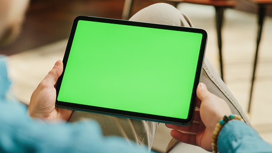 Young Man Scrolling and Tapping on Content on Tablet Computer with Green Screen Mock Up Display. Male Relaxing at Home, Reading Social Media Posts on Mobile Device. Close Up Over the Shoulder Photo.