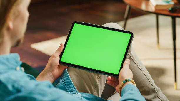istock Young Man Scrolling and Tapping on Content on Tablet Computer with Green Screen Mock Up Display. Male Relaxing at Home, Reading Social Media Posts on Mobile Device. Close Up Over the Shoulder Photo. 1447976623