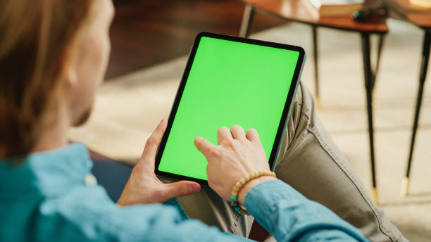 istock Young Man Scrolling Content on Tablet Computer with Green Screen Mock Up Display. Male Relaxing at Home, Reading Social Media Posts on Mobile Device. Close Up Over the Shoulder Photo. 1447976639