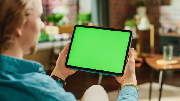istock Young Man Holding Tablet Computer with Green Screen Mock Up Display. Male Relaxing at Home, Watching Videos and Reading Social Media Posts on Mobile Device. Close Up Over the Shoulder Photo. 1447976679