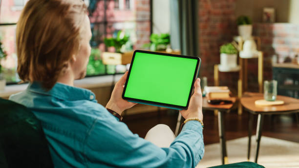 istock Young Man Holding Tablet Computer with Green Screen Mock Up Display. Male Relaxing at Home, Watching Videos and Reading Social Media Posts on Mobile Device. Close Up Over the Shoulder Photo. 1447976703