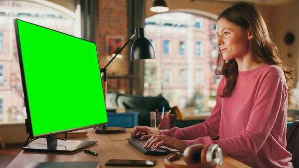 istock Young Pretty Female Working from Home on Desktop Computer with Green Screen Mock Up Display. Creative Woman Checking Social Media, Browsing Internet. Loft Apartment with City View from Big Window. 1447977747