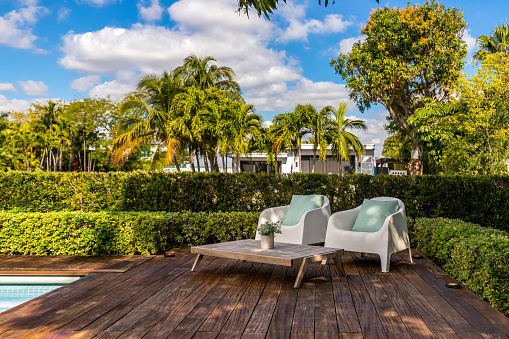 Beautiful patio with dark wood floor, swimming pool, bushes, trees, wooden coffee table lounge chairs, blue sky with clouds in Biscayne Point