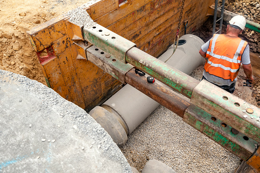 Builder installing big diameter concrete drainage pipe protected by trench support system during deep drainage works