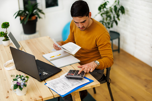 A young businessman at a home office is working and calculating business results using a calculator and the internet.