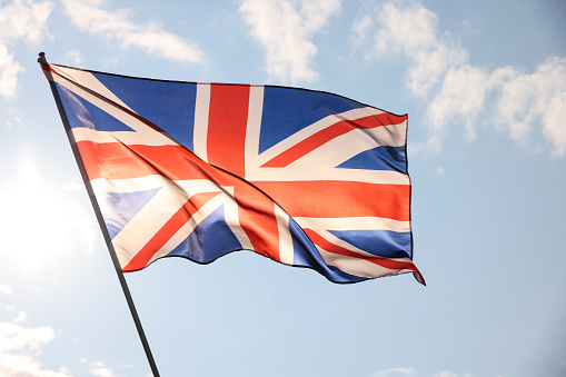 UK Great Britain national flag flying and waving in the wind, backlit in golden hour before sunset, over cloudy blue sky, symbol of British patriotism, low angle, side view