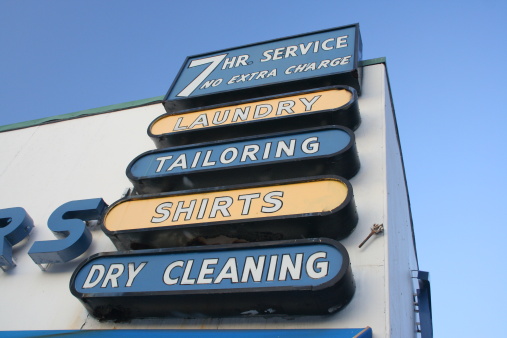 A multi-part laundry sign advertising their services, 7-hr service, no extra charge, laundry, tailoring, shirts, dry cleaning.  Blue & yellow sign against bold blue sky