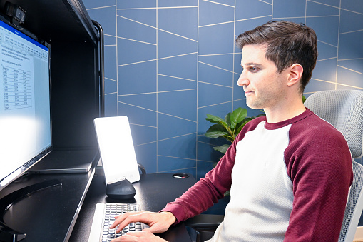 A young man is using a sunlight simulator lamp in his home office to combat the effects of Seasonal Affective Disorder in winter. The light is shining on his face and the sun lamp is positioned sideways as intended to emit 10,000 lux light at his eyes. The man is working on the computer while enjoying the benefits of improved mood. The home office has a blue geometric wallpaper in the background.