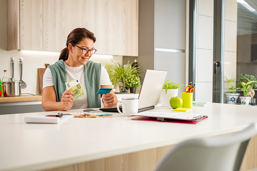 Hispanic smiling woman in kitchen using laptop and holding credit card and 100 Euro bill for online shopping and payments. High resolution 42Mp indoors digital capture taken with SONY A7rII and Zeiss Batis 40mm F2.0 CF lens