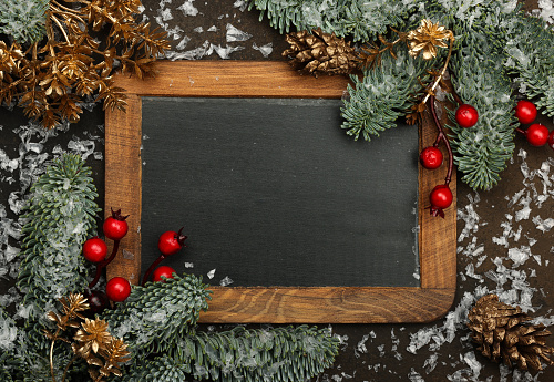Close up vintage black chalkboard sign with wooden frame and Christmas decoration of fresh spruce branches, golden cones, red holly berries and snow, copy space