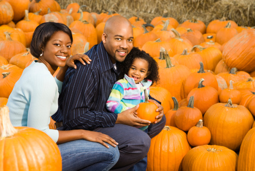 African American parents and daughter picking out pumpkin and smiling at outdoor market.