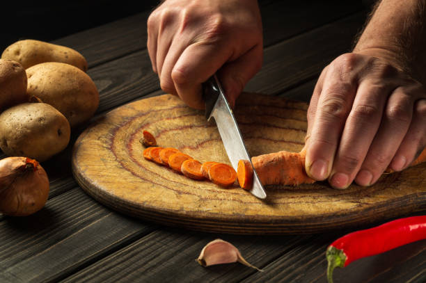 istock Slicing carrots on a wooden board for cooking. Chef hands with a knife while working on the kitchen table. Carrot diet 1448005029