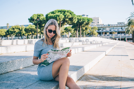 Blond female in casual clothes and sunglasses sitting on steps and reading map during trip in Barcelona City on sunny day