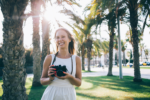 Joyful young female in white dress standing with photo camera in hands in city park with palm trees in sunny summer day in Barcelona