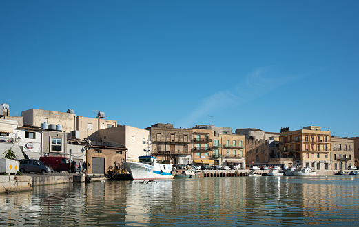 At the port of Mazara del Vallo there are many houses on the river against a blue sky. The houses are reflected in the water. Old boats are attached to the shore.