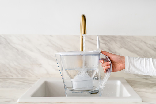 cropped shot of female hand hold pitcher with filter and pouring water from tap at kitchen on wall background with copy space, healthy lifestyle concept