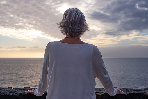 Rear view of senior woman standing in front of the sea at sunset looking at the horizon over the water. Peace, solitude, relaxation, meditation
