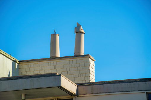 Chimney pipes with metal vents on the top of the house or home with square block style design with white stucco exterior. In the late afternoon sun and shade with blue and white gradient sky suburbs.