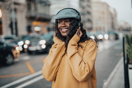 A young black woman, a happy black woman walks the streets of Barcelona, listening to music on wireless headphones, dressed casually and wearing a cap on her head