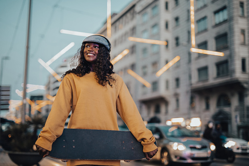 A young woman, a young black woman with a skateboard in her hands walks down the street of Barcelona, she is dressed hipster casually in a yellow sweater and has a cap on her head.