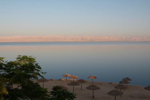 The rising sun illuminating the rocky desert coast across the Dead Sea, and just beginning to touch the tops of beach umbrellas on the near side.