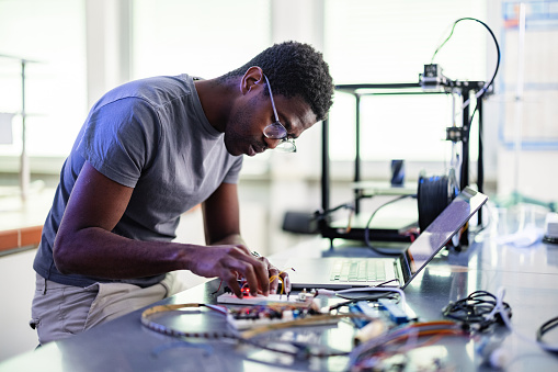 Young African-American technician sitting in the chair, thinking and repairing his 3D printer in the laboratory.