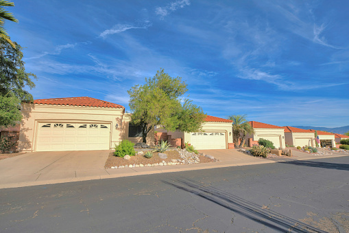 Attached garages in a subdivision at Tucson, Arizona. There is an asphalt road with cracks at the front of the driveway with white garage doors and concrete tile roofs against the blue sky.