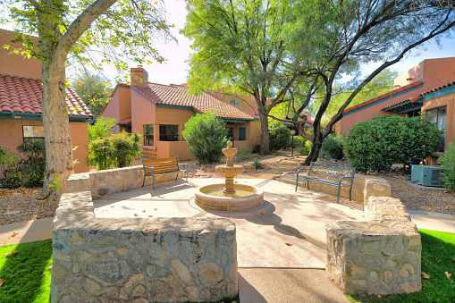 Small water fountain with two garden benches in the middle of mediterranean houses- Tucson, AZ. Outdoor lounge in a against the view of houses, trees, and a pathway on the right with lamp post.