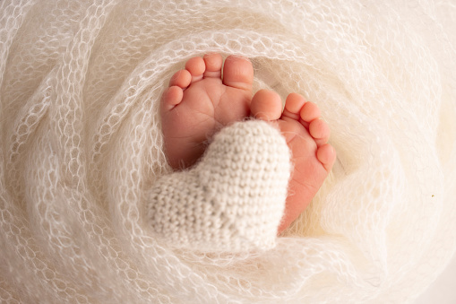 The tiny foot of a newborn baby. Soft feet of a new born in a white wool blanket. Close up of toes, heels and feet of a newborn. Knitted white heart in the legs of a baby. Macro photography.