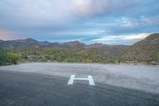 Helipad near the field of saguaro cactuses at Sabino Canyon State Park in Tucson, AZ. There is a view of mountain range against the cloudy sky from the helipad at the front.