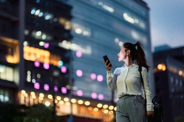 Young businesswoman using smartphone in financial district, confidence young woman using smart phone on her way off work outside office building at night woman trading stocks stock pictures, royalty-free photos & images
