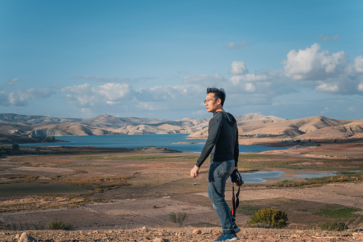 Asian Chinese man holding mirrorless camera walking at beautiful nature landscape near Fez, Morocco