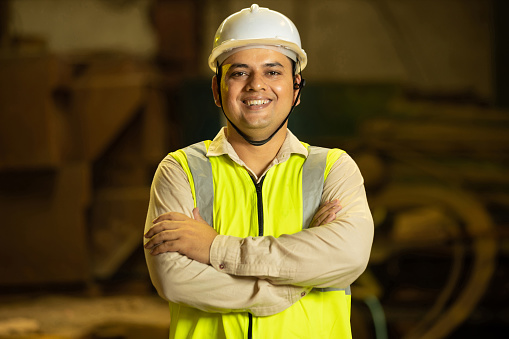 Indian Mechanical Engineer wearing safety yellow hard hat smiling while standing cross arm. Industrial factory worker concept.