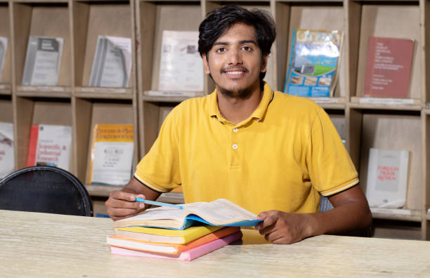 jeune étudiant indien lisant un livre étudiant dans la bibliothèque de l’université avec une étagère derrière. travailler sur une mission ou un projet. - book working college student classroom photos et images de collection