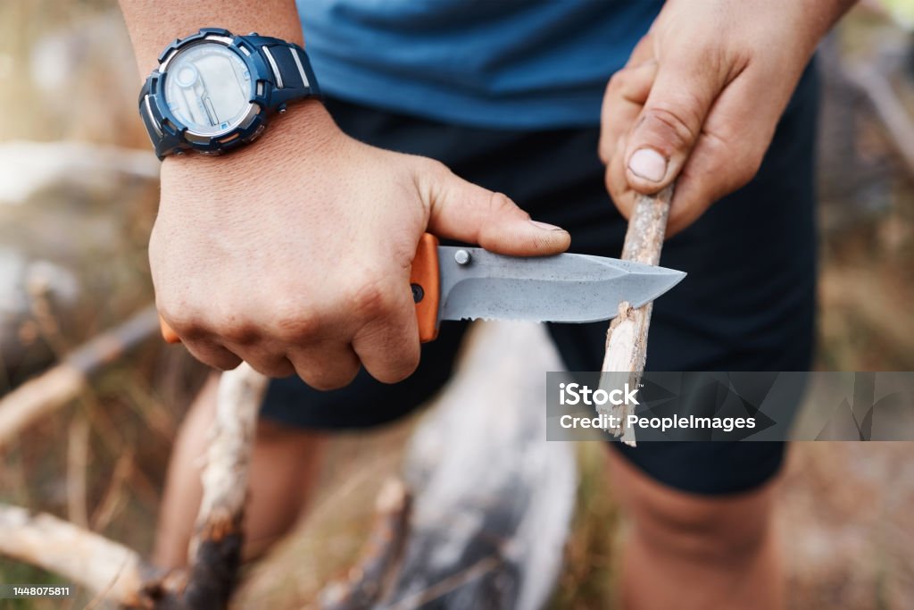 Hands, knife and cutting with a hiking man carving a stick outdoor in nature while camping for adventure. Wood, weapon and tool with a male camper slicing a twig in the mountains for survival Penknife Stock Photo