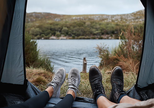 Shoes, camping and tent with a couple by a lake out in nature while hiking for adventure or travel together. Water, forest and freedom with a man and woman outdoor in the mountain to relax or getaway