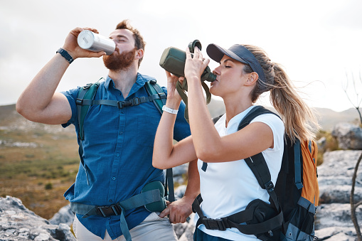 Water, hiking and couple trekking in the mountains for travel, peace and adventure during summer. Thirsty, tired and young man and woman with a drink from a bottle while walking in nature for holiday
