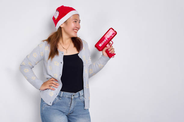 счастливая молодая азиатская женщина в рождественской шляпе и с денежным пистолетом, изолированная на белом фоне - guns and money стоковые фото и изображения