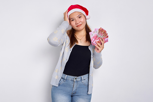 Smiling young Asian woman wearing Christmas hat holding rupiah banknotes for Christmas gift isolated on white background