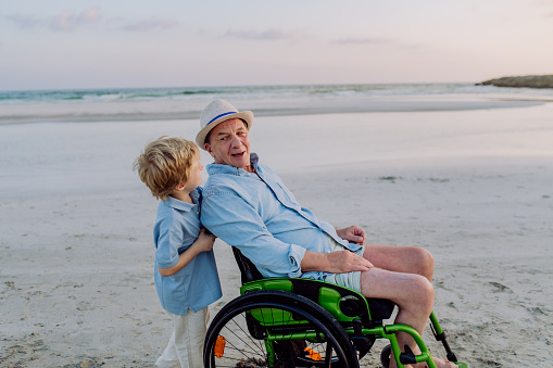 Little boy pushing his granfather on the wheelchair, enjoying sea together.