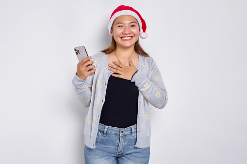 Happy young Asian woman in Christmas hat holding smartphone and holding chest isolated on white background