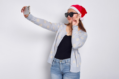 Excited young Asian woman in a Christmas hat doing a selfie shot on a mobile cell phone post a photo on social network isolated on white background