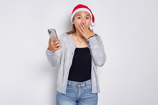 Shocked young Asian woman in a Christmas hat holding mobile phone, covering mouth with hand isolated on white background
