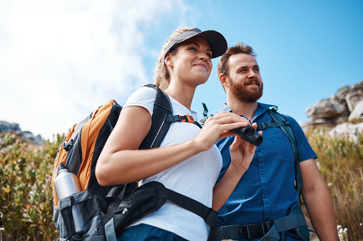 Hiking, binoculars and love with a couple on a mountain, enjoying a view while walking out in nature together. Summer, adventure and backpack with a man and woman sightseeing outdoor in the mountains