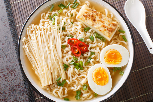 white ceramic bowl of ramen soup is filled with enoki mushrooms, noodles, green onions, tofu and soft boiled eggs close-up on the table. Horizontal top view from above